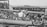A crowded deck of a ship features numerous men dressed in military uniforms, some of whom are sitting and others standing. Two of these men are engaged in what appears to be a task or activity involving a large piece of machinery or equipment. The sea is visible in the background, indicating that the ship is at sea.