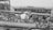 A crowded deck of a ship features numerous men dressed in military uniforms, some of whom are sitting and others standing. Two of these men are engaged in what appears to be a task or activity involving a large piece of machinery or equipment. The sea is visible in the background, indicating that the ship is at sea.
