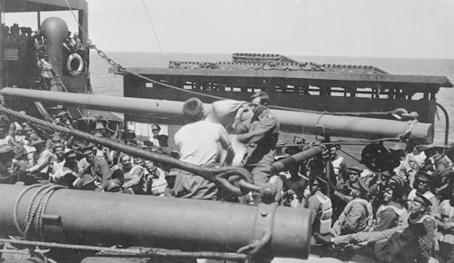 A crowded deck of a ship features numerous men dressed in military uniforms, some of whom are sitting and others standing. Two of these men are engaged in what appears to be a task or activity involving a large piece of machinery or equipment. The sea is visible in the background, indicating that the ship is at sea.