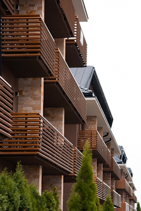 Several modern balconies with wooden railings fixed to a building with a stone facade. The building has a series of neatly arranged balconies, creating a repeating pattern. In the foreground, green shrubs add a natural element to the otherwise architectural scene.
