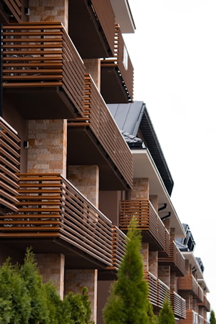 Several modern balconies with wooden railings fixed to a building with a stone facade. The building has a series of neatly arranged balconies, creating a repeating pattern. In the foreground, green shrubs add a natural element to the otherwise architectural scene.