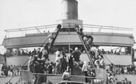 A large crowd of people climbs two narrow gangways leading to a higher platform on a ship, with many others gathered below and on the upper deck. The focus is on the human activity and interaction as people move among each other in an orderly fashion.