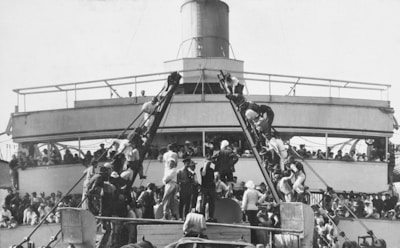 A large crowd of people climbs two narrow gangways leading to a higher platform on a ship, with many others gathered below and on the upper deck. The focus is on the human activity and interaction as people move among each other in an orderly fashion.