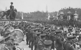 A historic military parade with soldiers in uniform marching and playing musical instruments. A large crowd gathers, observing the proceedings in an urban setting featuring a statue and elaborate gates.
