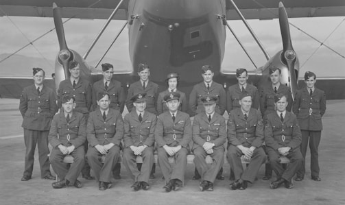 A group of fifteen men in military uniforms are posed in front of a large aircraft. They are arranged in two rows, with the front row seated and the back row standing. The aircraft's propeller and structure are visible behind them on an overcast day.