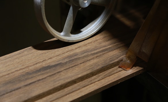 A metallic wheel is positioned next to a wooden surface with visible grain patterns. To the right, there's a rusted metal piece resting on the wood, casting a shadow due to directional lighting.