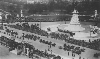 A large military procession is taking place around a grand, ornate monument in a city square. The scene includes rows of soldiers marching, horse-drawn carriages, and a large crowd of spectators surrounding the area.