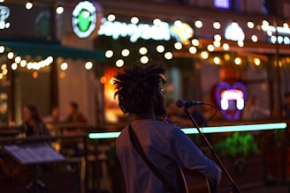 A musician with dreadlocks is performing outdoors at night, standing in front of a microphone and a guitar. The scene has decorative string lights, creating a warm and inviting atmosphere. People are seated at the background in a café or bar setting, enjoying the evening.