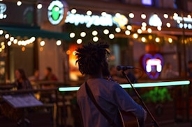 A musician with dreadlocks is performing outdoors at night, standing in front of a microphone and a guitar. The scene has decorative string lights, creating a warm and inviting atmosphere. People are seated at the background in a café or bar setting, enjoying the evening.