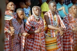 A group of people dressed in traditional Maasai attire, featuring red and white checkered patterns, are engaged in a cultural or ceremonial activity. Some individuals are holding sticks, and others are adorned with beadwork. The scene is lively with a backdrop of onlookers.