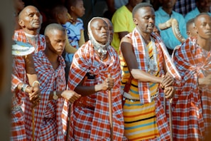 A group of people dressed in traditional Maasai attire, featuring red and white checkered patterns, are engaged in a cultural or ceremonial activity. Some individuals are holding sticks, and others are adorned with beadwork. The scene is lively with a backdrop of onlookers.