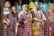 A group of people dressed in traditional Maasai attire, featuring red and white checkered patterns, are engaged in a cultural or ceremonial activity. Some individuals are holding sticks, and others are adorned with beadwork. The scene is lively with a backdrop of onlookers.