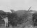 A soldier, wearing a helmet and military gear, is positioned in a trench fortified with sandbags, aiming an anti-aircraft gun into the sky. The trench is surrounded by a rugged landscape, with makeshift fencing and overgrown vegetation. In the distance, the remnants of a crashed or damaged military aircraft can be seen protruding.