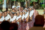 A group of people dressed in traditional attire, adorned with colorful garments and intricate beadwork around their necks. They appear to be participating in a cultural or ceremonial event, standing closely together in a formation. The scene is set outdoors with blurred greenery in the background, indicating a bright, sunny day.
