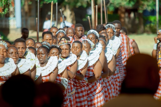 A group of people dressed in traditional attire, adorned with colorful garments and intricate beadwork around their necks. They appear to be participating in a cultural or ceremonial event, standing closely together in a formation. The scene is set outdoors with blurred greenery in the background, indicating a bright, sunny day.