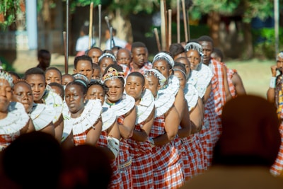 A group of people dressed in traditional attire, adorned with colorful garments and intricate beadwork around their necks. They appear to be participating in a cultural or ceremonial event, standing closely together in a formation. The scene is set outdoors with blurred greenery in the background, indicating a bright, sunny day.