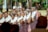 A group of people dressed in traditional attire, adorned with colorful garments and intricate beadwork around their necks. They appear to be participating in a cultural or ceremonial event, standing closely together in a formation. The scene is set outdoors with blurred greenery in the background, indicating a bright, sunny day.