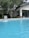 A clear blue swimming pool is situated in the foreground with water flowing into it from a fountain. The pool is surrounded by tall pine trees and a modern house with large glass windows and a sloping roof is visible in the background.