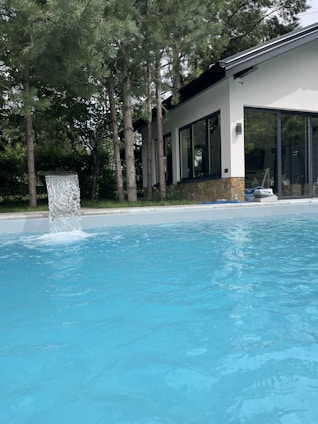 A clear blue swimming pool is situated in the foreground with water flowing into it from a fountain. The pool is surrounded by tall pine trees and a modern house with large glass windows and a sloping roof is visible in the background.