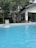 A clear blue swimming pool is situated in the foreground with water flowing into it from a fountain. The pool is surrounded by tall pine trees and a modern house with large glass windows and a sloping roof is visible in the background.