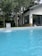 A clear blue swimming pool is situated in the foreground with water flowing into it from a fountain. The pool is surrounded by tall pine trees and a modern house with large glass windows and a sloping roof is visible in the background.