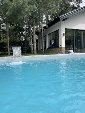 A clear blue swimming pool is situated in the foreground with water flowing into it from a fountain. The pool is surrounded by tall pine trees and a modern house with large glass windows and a sloping roof is visible in the background.