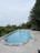 A rectangular swimming pool with clear blue water is surrounded by a tiled patio. Tall pine trees line the right side, providing a natural, lush backdrop. The pool has a step entry, and railings are present on the left side. In the distance, a variety of trees and shrubs add to the greenery, creating a serene and secluded atmosphere.