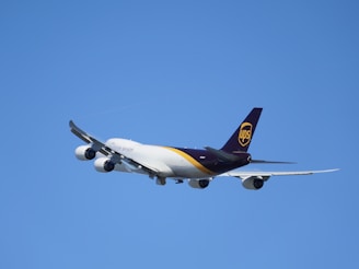 A large cargo airplane with a distinctive white and dark brown color scheme and a UPS logo on the tail is flying against a clear blue sky. The aircraft is seen from a rear-side angle, showcasing its four engines and wide wingspan.