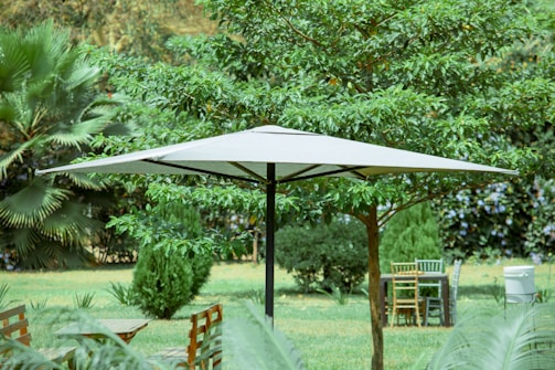 A large white umbrella providing shade in a lush garden setting. Surrounding the umbrella are various green trees and bushes, and there are wooden chairs and a table nearby. The scene is serene and suggests a well-maintained outdoor space.