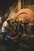 A person engaged in sculpting clay figures, surrounded by partially completed statues. The workspace is cluttered with materials and tools, and the environment appears rustic with dim lighting.