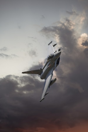 A fighter jet ascending through a dynamic sky filled with dark, dramatic clouds with hints of sunlight breaking through.