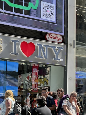 A large 'I Love NY' sign is displayed on the exterior of a building. Below it, several people are walking and standing on the street. Inside the store, mannequins and various products can be seen through the glass windows. A colorful QR code and a digital screen are also visible on the building.
