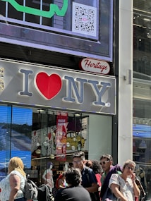 A large 'I Love NY' sign is displayed on the exterior of a building. Below it, several people are walking and standing on the street. Inside the store, mannequins and various products can be seen through the glass windows. A colorful QR code and a digital screen are also visible on the building.