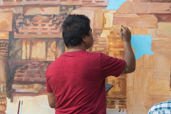 A person wearing a red shirt is painting a detailed mural of architectural structures, likely an ancient or historical building. The person is facing away from the camera and holding a paintbrush in their right hand. The mural features intricate details and a mix of warm colors, with some parts of the mural still unpainted or light blue.