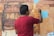 A person wearing a red shirt is painting a detailed mural of architectural structures, likely an ancient or historical building. The person is facing away from the camera and holding a paintbrush in their right hand. The mural features intricate details and a mix of warm colors, with some parts of the mural still unpainted or light blue.