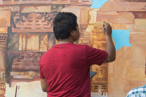 A person wearing a red shirt is painting a detailed mural of architectural structures, likely an ancient or historical building. The person is facing away from the camera and holding a paintbrush in their right hand. The mural features intricate details and a mix of warm colors, with some parts of the mural still unpainted or light blue.