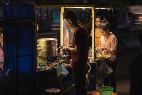 A street food vendor is preparing food at a small stall illuminated by warm lighting. Two women are standing nearby, watching or waiting for their order. The setup includes a steaming pot, various utensils, and ingredients scattered on the counter. The scene takes place at night with a dimly lit street in the background.