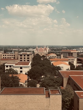 Aerial view of an urban area featuring a prominent building with distinct architectural features, such as pointed rooftops. Surrounding the central structure are multiple rectangular buildings with flat roofs and red tiling. A line of trees leads the eye toward the focal point, while patches of greenery surround the area. The distant horizon and cloudy sky create a serene backdrop.