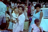 A crowded marketplace scene with multiple people. A woman is in the center holding a baby. Another woman in a blue dress stands nearby looking towards the camera. A child is in the foreground eating something. Various people can be seen in the background, along with market stalls and a car.