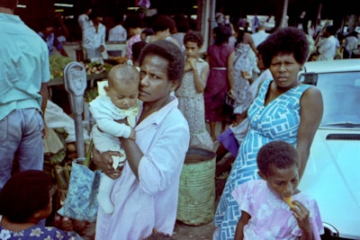 A crowded marketplace scene with multiple people. A woman is in the center holding a baby. Another woman in a blue dress stands nearby looking towards the camera. A child is in the foreground eating something. Various people can be seen in the background, along with market stalls and a car.
