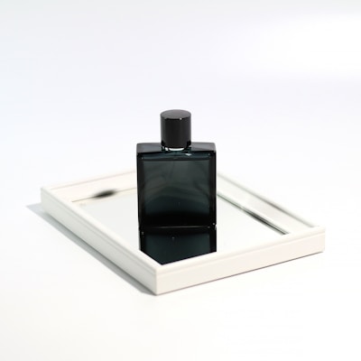 A sleek, dark-colored perfume bottle is placed upright on a rectangular mirror. The mirror is framed with a thin, white border, and reflects part of the bottle's base and cap, creating an elegant and minimalist presentation. The background is a soft white, enhancing the focus on the reflective surface and bottle.
