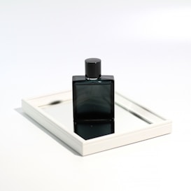 A sleek, dark-colored perfume bottle is placed upright on a rectangular mirror. The mirror is framed with a thin, white border, and reflects part of the bottle's base and cap, creating an elegant and minimalist presentation. The background is a soft white, enhancing the focus on the reflective surface and bottle.