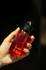 A hand is holding a vibrant red perfume bottle with a black cap. The background is dark, which highlights the glossy finish of the bottle.