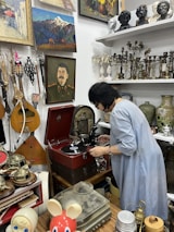 A person is adjusting a vintage record player in what appears to be an antique shop or collection room. The walls are adorned with various paintings, including landscapes and portraits. Shelves are filled with classical busts, candlesticks, and vases. Musical instruments like guitars are visible, as well as decorative objects, such as a red toy with large eyes in the foreground.