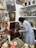 A person is adjusting a vintage record player in what appears to be an antique shop or collection room. The walls are adorned with various paintings, including landscapes and portraits. Shelves are filled with classical busts, candlesticks, and vases. Musical instruments like guitars are visible, as well as decorative objects, such as a red toy with large eyes in the foreground.