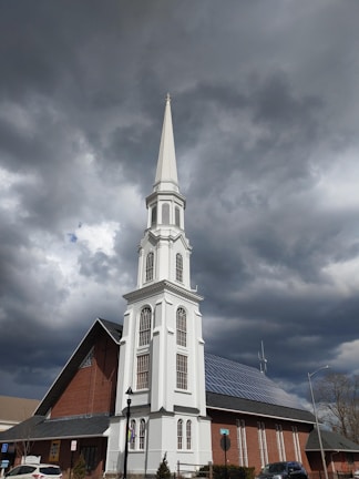 A tall, white steeple of a church with multiple windows rises against a backdrop of dark, cloudy skies. The church building features red brick walls, a gabled roof with solar panels, and a signboard near the entrance. A lamp post and some street signs are also visible in the surrounding area.