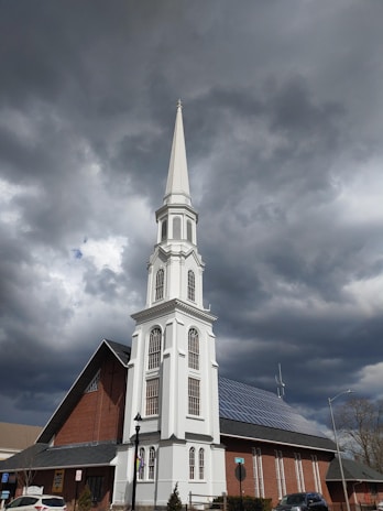 A tall, white steeple of a church with multiple windows rises against a backdrop of dark, cloudy skies. The church building features red brick walls, a gabled roof with solar panels, and a signboard near the entrance. A lamp post and some street signs are also visible in the surrounding area.