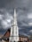 A tall, white steeple of a church with multiple windows rises against a backdrop of dark, cloudy skies. The church building features red brick walls, a gabled roof with solar panels, and a signboard near the entrance. A lamp post and some street signs are also visible in the surrounding area.