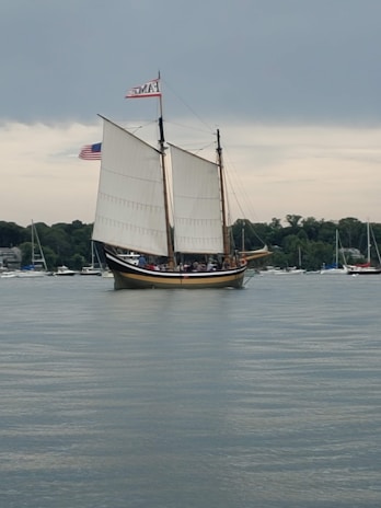 A large sailboat with two prominent sails glides across a calm body of water. Several people are visible on the deck, and the boat features a small American flag alongside another flag. In the background, there are numerous smaller boats anchored near a tree-lined shore under a cloudy sky.