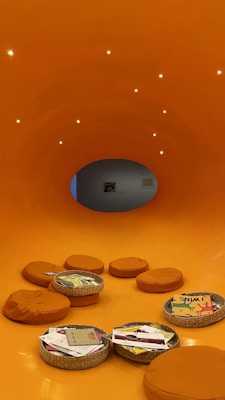 A vibrant orange room with circular padded seats and woven baskets scattered on the floor. The baskets contain various illustrated books and papers. Soft lighting gives the room a cozy atmosphere. The orange hue of the walls and floor creates an inviting and warm environment. A circular opening at the back leads to another room.